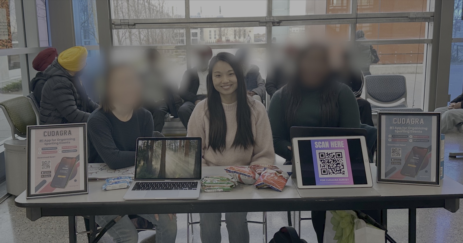 Sitting at a table in the Genesis Centre lobby with a laptop, snacks, a QR code sign-up on a tablet, and Cudagra posters, faces of passersby blurred